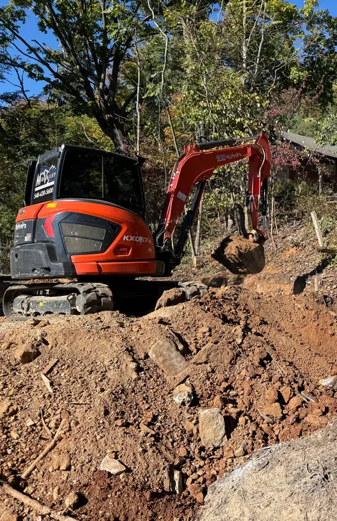 Mid Valley Grounds Kubota KX057 excavator with the company logo and phone number on the cab, digging into a wooded slope.