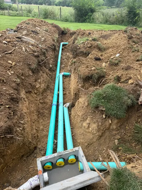 Two green sewer lines running the length of a deep trench across a grass field toward a distribution box.